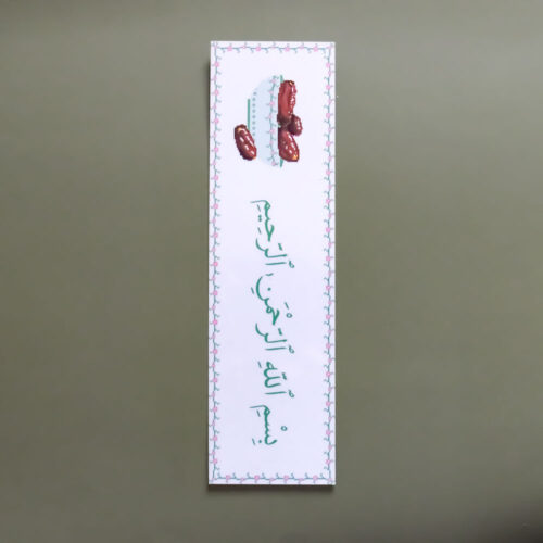 A close-up, top-down photograph shows a long, rectangular white bookmark with a vertical orientation resting on a solid olive-green surface. The bookmark features a thin decorative border of small pink and blue floral motifs. At the top, there is an illustration of a light blue bowl filled with reddish-brown dates. Below the bowl, the Arabic phrase "Bismillah ar-Rahman ar-Rahim" (In the name of Allah, the Most Gracious, the Most Merciful) is written in a graceful green script.