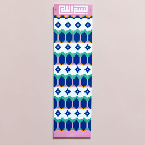 The image shows a long, rectangular bookmark with a vertical layout, resting on a solid grey surface. The design is divided into a dark blue top section featuring a pink mosque-style silhouette with a small crescent on top, set against a background of white stars. Below this, a series of decorative geometric borders in white, orange, and teal separate the top illustration from the main body. The rest of the bookmark consists of a light pink background covered in an all-over repeating pattern of white geometric, oval-like motifs.