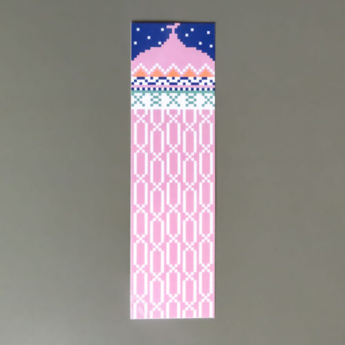 The image shows a long, rectangular bookmark with a vertical layout, resting on a solid grey surface. The design is divided into a dark blue top section featuring a pink mosque-style silhouette with a small crescent on top, set against a background of white stars. Below this, a series of decorative geometric borders in white, orange, and teal separate the top illustration from the main body. The rest of the bookmark consists of a light pink background covered in an all-over repeating pattern of white geometric, oval-like motifs.